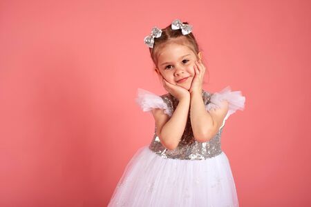 Portrait of cute little girl wearing white-pink dress, smiling and propping up her head with hands over pink backgroundの写真素材