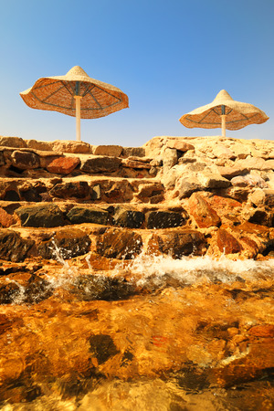 Summer landscape - the wave is breaking on the stone steps leading to the beach with wicker umbrellasの写真素材