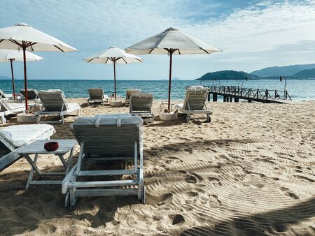 White soft sun loungers on the shore of a sandy sea beach. Tropical resort, morning sun, white clouds in the blue sky, view of the sea, mountains and the city in the backgroundの写真素材