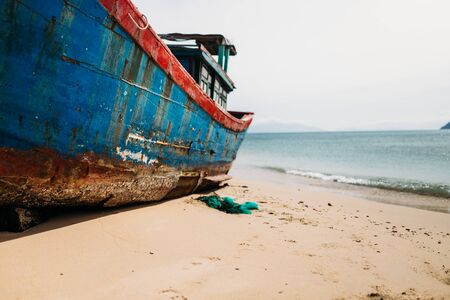 A traditional Asian fishing boat stands in a calm bay on the sandy shore. Old fishing boat, rust on the hull, White clouds on a blue sky. Travel in Vietnamの写真素材