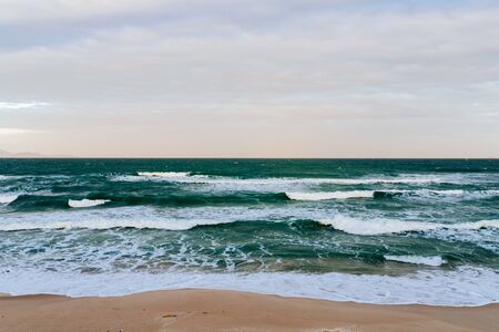 Sandy seashore, calm soft waves at sea, sunny weather, blue sky with white clouds. Empty long beach with a beautiful sea view. White foam from a sea waveの写真素材