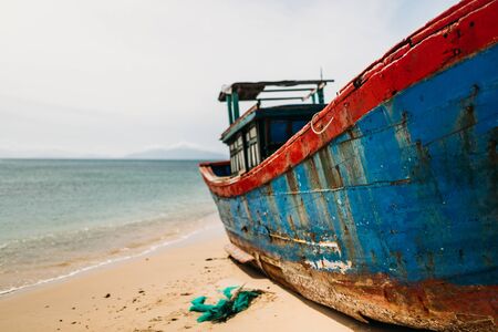 A traditional Asian fishing boat stands in a calm bay on the sandy shore. Old fishing boat, rust on the hull, White clouds on a blue sky. Travel in Vietnamの写真素材