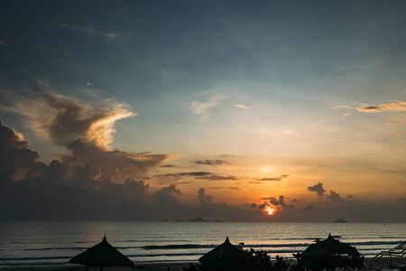 Beautiful sunrise, sunset of the sun through the clouds, the rays of the sun through the clouds, sandy seashore with small waves, warm morning light in a tropical area near the sea.の写真素材