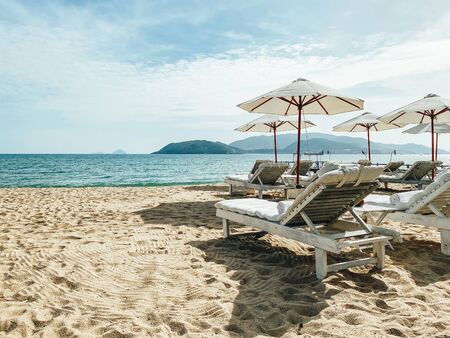 White soft sun loungers on the shore of a sandy sea beach. Tropical resort, morning sun, white clouds in the blue sky, view of the sea, mountains and the city in the backgroundの写真素材