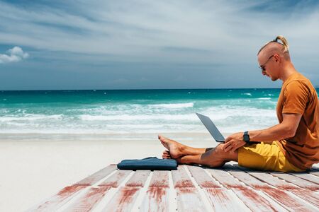 Young guy with long hair by the sea on a sunny day, working using his laptop, working remotely with a business. Bloner, freelancer, work through the Internet, work travel. Summer vacation. Sandy beach by the ocean with a wooden path. Copy placeの写真素材