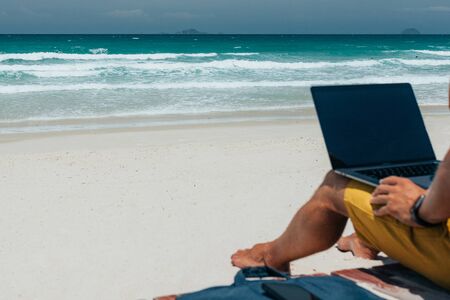 Young guy with long hair by the sea on a sunny day, working using his laptop, working remotely with...