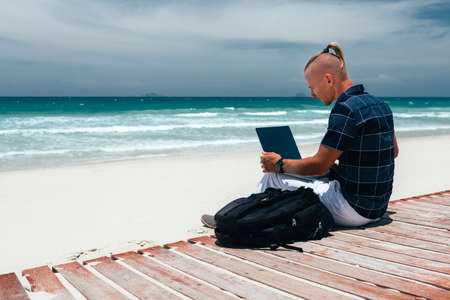 guy working on remote work on a journey using a laptop, sitting on the seashore on a sandy beach. Businessman working on remote work on a journeyの写真素材