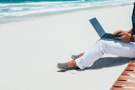 guy working using a laptop, sitting on the seashore on a sandy beach. working on remote work on a journey. Blogger, freelancer makes a content work planの写真素材