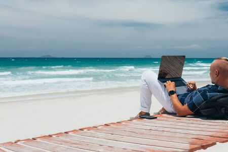 Blogger guy working using a laptop, on the seashore on a sandy beach. Businessman working on remote work on a journey. freelancer makes a content work planの写真素材