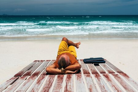 Young guy with long hair by the sea on a sunny day, working using his laptop, working remotely with a business. Bloner, freelancer, work through the Internet, work travel. Summer vacation. Sandy beach by the ocean with a wooden path. Copy placeの写真素材