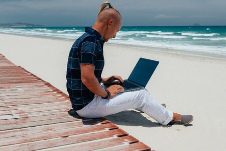 Young guy working using a laptop, sitting on the seashore on a sandy beach. Businessman working on remote work on a journey. Blogger, freelancer makes a contentの写真素材