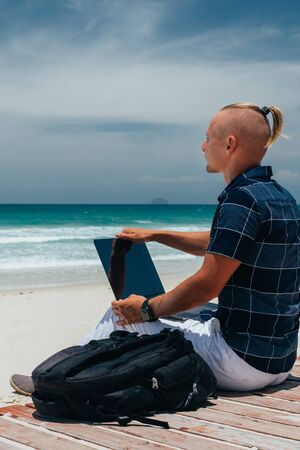 Young guy Looks at the sea side working using a laptop, sitting on the seashore on a sandy beach. Businessman on remote work on a journey. Blogger, freelancer makes a content workの写真素材