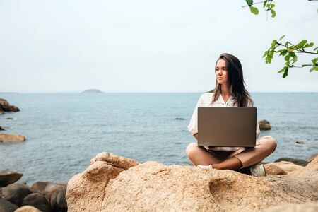 Young successful girl working using a laptop, sitting on a rocky seashore. Business woman working on remote work while traveling, blogger, freelancer, compiles content plan, work online. copy spaceの写真素材