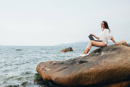 Young successful, smiling girl working using a laptop, sitting on a rocky seashore. Business woman working on remote work while traveling, blogger, work online. copy spaceの写真素材