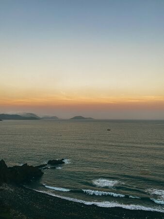 Sunset on the sea with a panoramic view from the top of the cliff. View of the rocky sea shore, waves on the shore. Mountains on the horizon. Beautiful sky color from the setting sun. Beautiful dramatic seascapeの写真素材