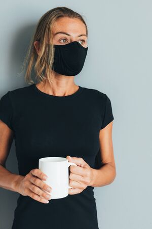 Portrait of a young woman in a black mask with a cup of coffee in hand, during isolation, stay at home, look to the side, a clean background wall with space for text, copy spaceの写真素材