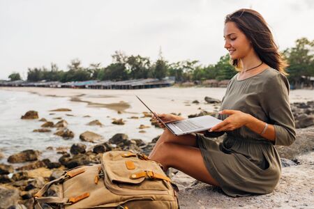 Young woman blogger working remotely with a laptop using the internet, on the seashore, sitting on the beach. Work on vacation while traveling. Looking at the monitor. Freelance concept. Copy placeの写真素材