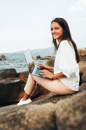 Young successful, smiling girl working using a laptop, sitting on a rocky seashore. Business woman working on remote work while traveling, blogger, freelancer, compiles content plan, work online. copy spaceの写真素材