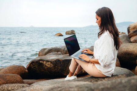 Young girl working using a laptop, sitting on a rocky seashore. Business woman working on remote work while traveling, blogger, freelancer, compiles content plan, work online. copy spaceの写真素材