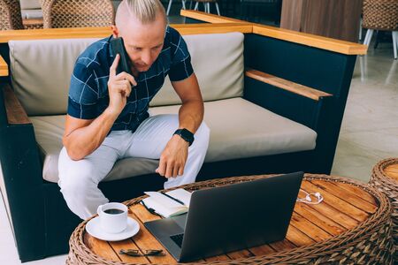 A male young businessman, doing remote work while sitting in a cafe with a cup of coffee in the morning. Uses modern technology. A laptop, Phones to clarify information. Successful computer work on the Internetの写真素材