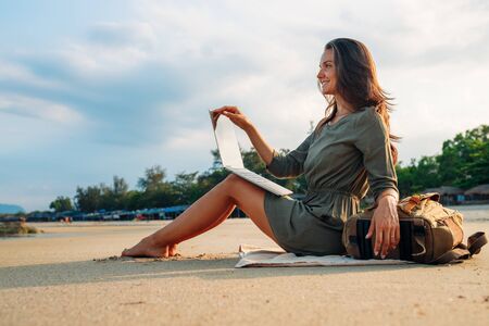 A young girl works on a laptop at sea. Work as a freelancer away from the office. Work while traveling. Successful business lady. Modern technology for work. Internet work, Sitting on the beach with a backpackの写真素材