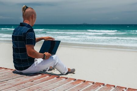 Young guy Open a laptop for work, sitting on the seashore on a sandy beach. Businessman working on remote work on a journey. Blogger, freelancer makes a content work planの写真素材