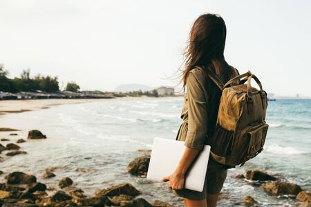 A view from the back of a girl on a rocky seashore, a traveler. Freelance worker with a backpack on his shoulders and with a laptop in his hands, Sea view, concept of travel, vacation. Place for text, copy placeの写真素材