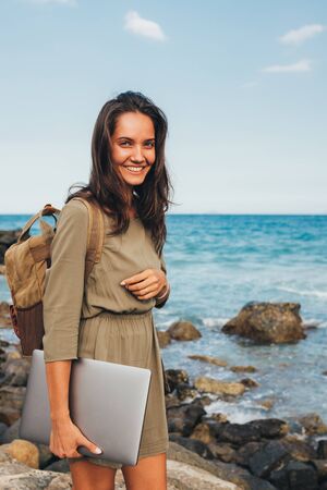 Freelancer  Girl on a stone pier with a laptop in her hands and a brown backpack on the bedroom. Smiling at camera, concept of distant work, freelance, sea voyage, vacation. Copy placeの写真素材