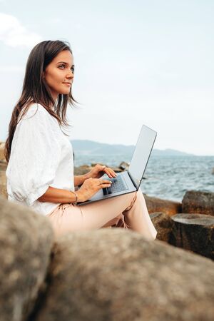 Young girl working using a laptop, sitting on a rocky seashore. Business woman working on remote work while traveling, freelancer, work online. copy spaceの写真素材