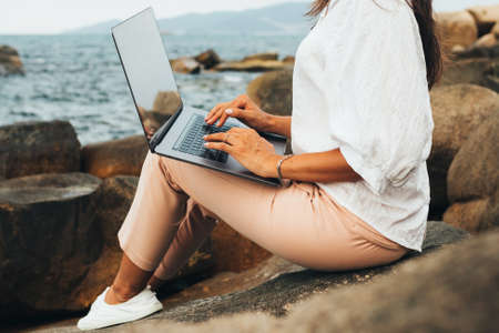 girl working using a laptop, sitting on a rocky seashore. Business woman working on remote work while traveling, blogger, freelancer, compiles content plan, work online. copy spaceの写真素材