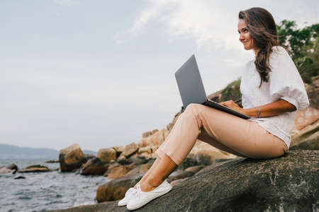 Young, smiling girl working using a laptop, sitting on a rocky seashore. Business woman working on remote work while traveling, freelancer, compiles content plan, work online. copy spaceの写真素材