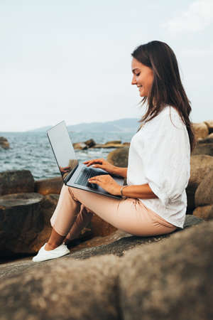 Young successful, smiling girl working using a laptop, sitting on a rocky seashore. Business woman working on remote work while traveling, blogger, freelancer, compiles content plan, work online. copy spaceの写真素材
