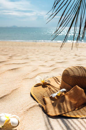 Straw hat with frangipani flowers on white sea sand. With beautiful seascape, the blue sea in the background, Palm leaf shadow. Copy space, background for advertisement of a seaside vacationの写真素材
