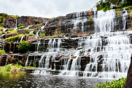 Beautiful, wide waterfall in a popular tourist spot. A powerful stream of water along rocky ledges, cascading into a lake powdered with greenery. Long exposure shootingの写真素材