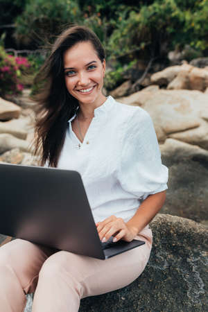 Young successful, smiling girl working using a laptop, sitting on a rocky seashore. Business woman working on remote work while traveling, blogger, freelancer, compiles content plan, work online. copy spaceの写真素材