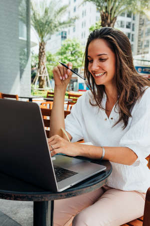 Freelance concept. Attractive girl uses her laptop while sitting in a cafe on the street. looks through mail on a laptop and makes notes in a notebook. Tropical palms in the backgroundの写真素材