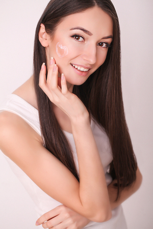 beauty, skin care and people concept - smiling young woman applying cream to face and looking to mirror at home bathroom.の写真素材