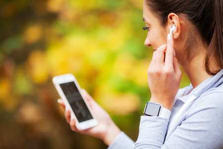 Photo of Joyful Fitness Woman 30s in Sportswear Touching Earpod and Holding Mobile Phone, While Resting in Green Parkの写真素材
