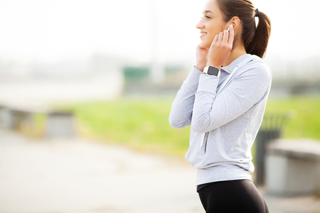 Photo of Joyful Fitness Woman 30s in Sportswear Touching Earpod and Holding Mobile Phone, While Resting in Green Parkの写真素材