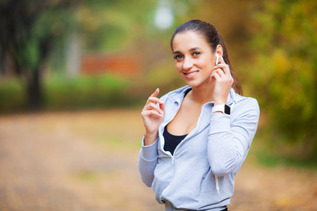 Photo of Joyful Fitness Woman 30s in Sportswear Touching Earpod and Holding Mobile Phone, While Resting in Green Parkの写真素材