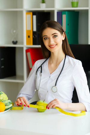 Health. Diet and Healthy Nutrition. Doctor Dietitian Holding in Hands Fresh Green Apple and Centimeter Ribbon. Smiles Beautiful and Young Doctor in the Light Room. High Resolutionの写真素材