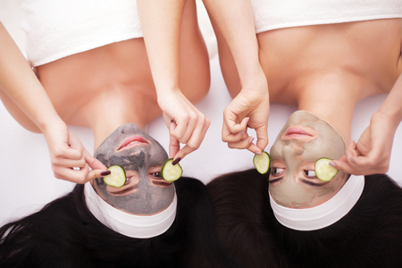 Home spa. Two beautiful young women holding pieces of cucumber on their eyes and smiling while on the bedの写真素材