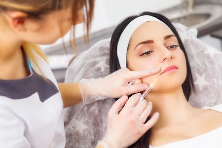 Close up of hands of cosmetologist making botox injection in female lips. She is holding syringe. The young beautiful woman is receiving procedure with enjoymentの写真素材