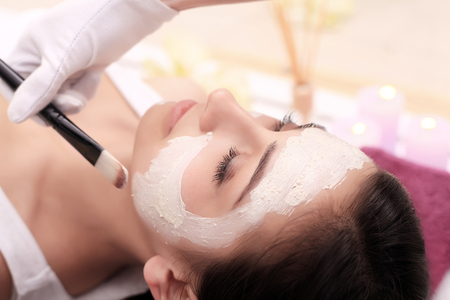 Beautiful young woman is getting facial clay mask at spa, lying with cucumbers on eyesの写真素材