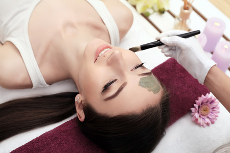 Beautiful young woman is getting facial clay mask at spa, lying with cucumbers on eyesの写真素材