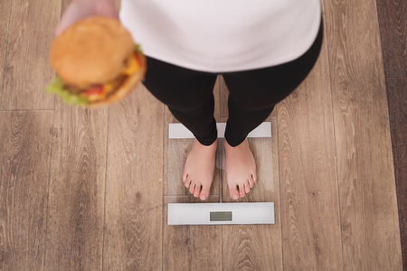 Diet And Fast Food Concept. Overweight Woman Standing On Weighing Scale Holding Burger Hamburger . Unhealthy Junk Food. Dieting, Lifestyle. Weight Loss. Obesity. Top Viewの写真素材