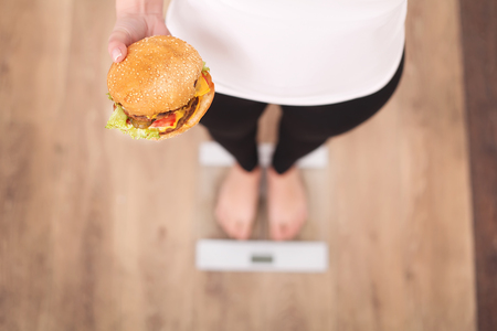 Diet And Fast Food Concept. Overweight Woman Standing On Weighing Scale Holding Burger ( Hamburger ). Unhealthy Junk Food. Dieting, Lifestyle. Weight Loss. Obesity. Top Viewの写真素材