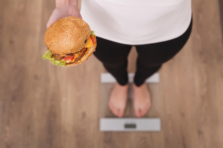 Diet And Fast Food Concept. Overweight Woman Standing On Weighing Scale Holding Burger Hamburger . Unhealthy Junk Food. Dieting, Lifestyle. Weight Loss. Obesity. Top Viewの写真素材
