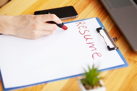 Job search. Female hand writes resume with lipstick on white sheet of paper. Wooden office desk with laptop, smartphone and suppliesの写真素材