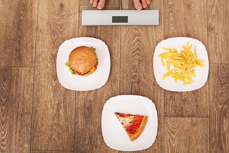 A young beautiful woman is on the scales. To choose between potato chips, pizza and burgers. The concept of healthy eating. Healthy Lifestyle. Diet.の写真素材
